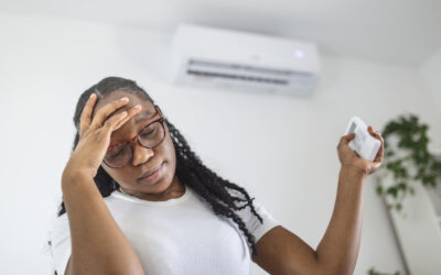 A woman in a white t-shirt and glasses rests her head on her hand in frustration as she uses her AC remote to try to adjust the temperature of the AC on the wall behind her.