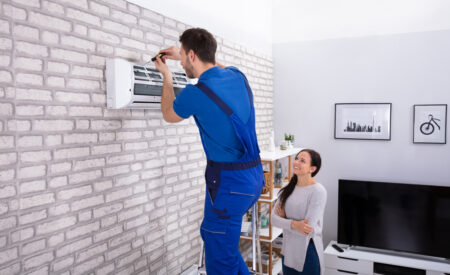 A Repairman In A Blue Uniform Installs A New Air Conditioner On A Brick Wall While Being Observed By A Female Homeowner In A White Sweater.