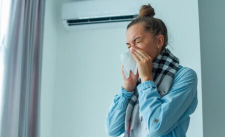 A Woman With Blonde Hair Tied Up Into A Bun Experiences Allergy Symptoms, Blowing Her Nose Into Tissue Due To Her Home’s Poor Indoor Air Quality From Her AC Unit.