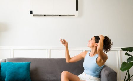 A woman appears distressed while sitting on a couch beside an air conditioner, pondering its role in reducing humidity.
