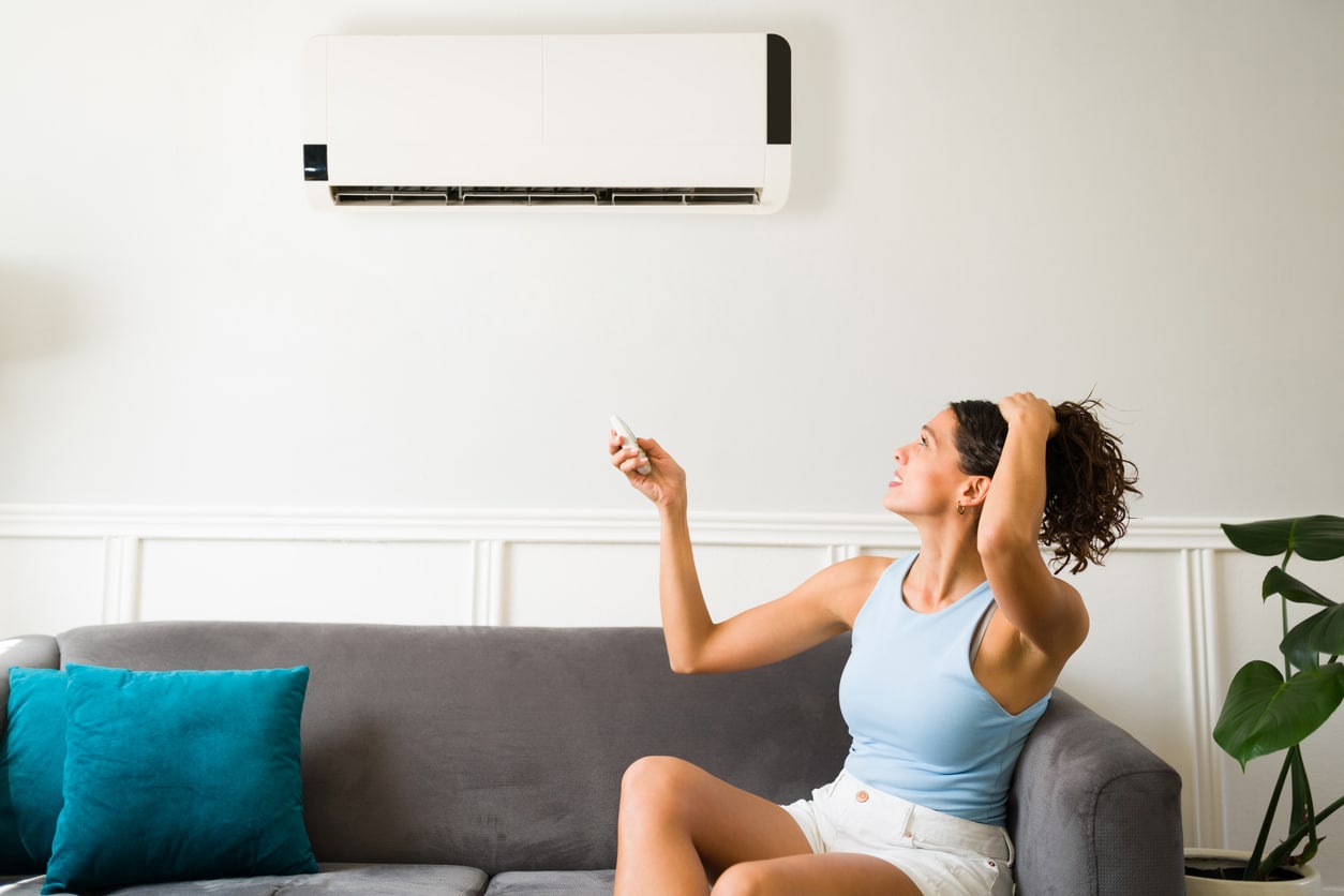 A woman appears distressed while sitting on a couch beside an air conditioner, pondering its role in reducing humidity.