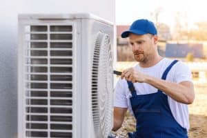 An HVAC technician wearing safety gear uses a power drill to replace an outdoor air conditioning unit.