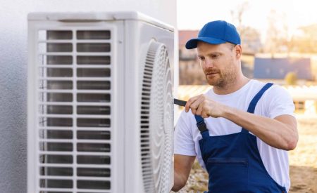 An HVAC technician wearing safety gear uses a power drill to replace an outdoor air conditioning unit.