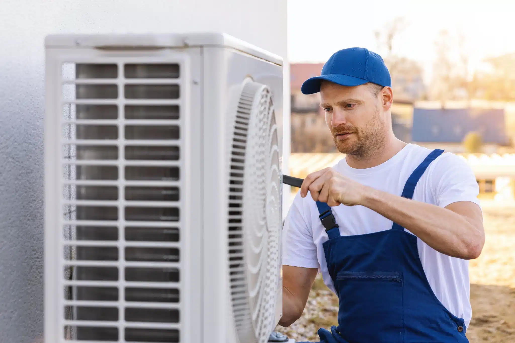 An HVAC technician wearing safety gear uses a power drill to replace an outdoor air conditioning unit.