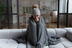 A woman wrapped in a blanket on a couch attempts to stay warm after her HVAC system struggles during a winter cold snap.