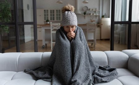 A woman wrapped in a blanket on a couch attempts to stay warm after her HVAC system struggles during a winter cold snap.