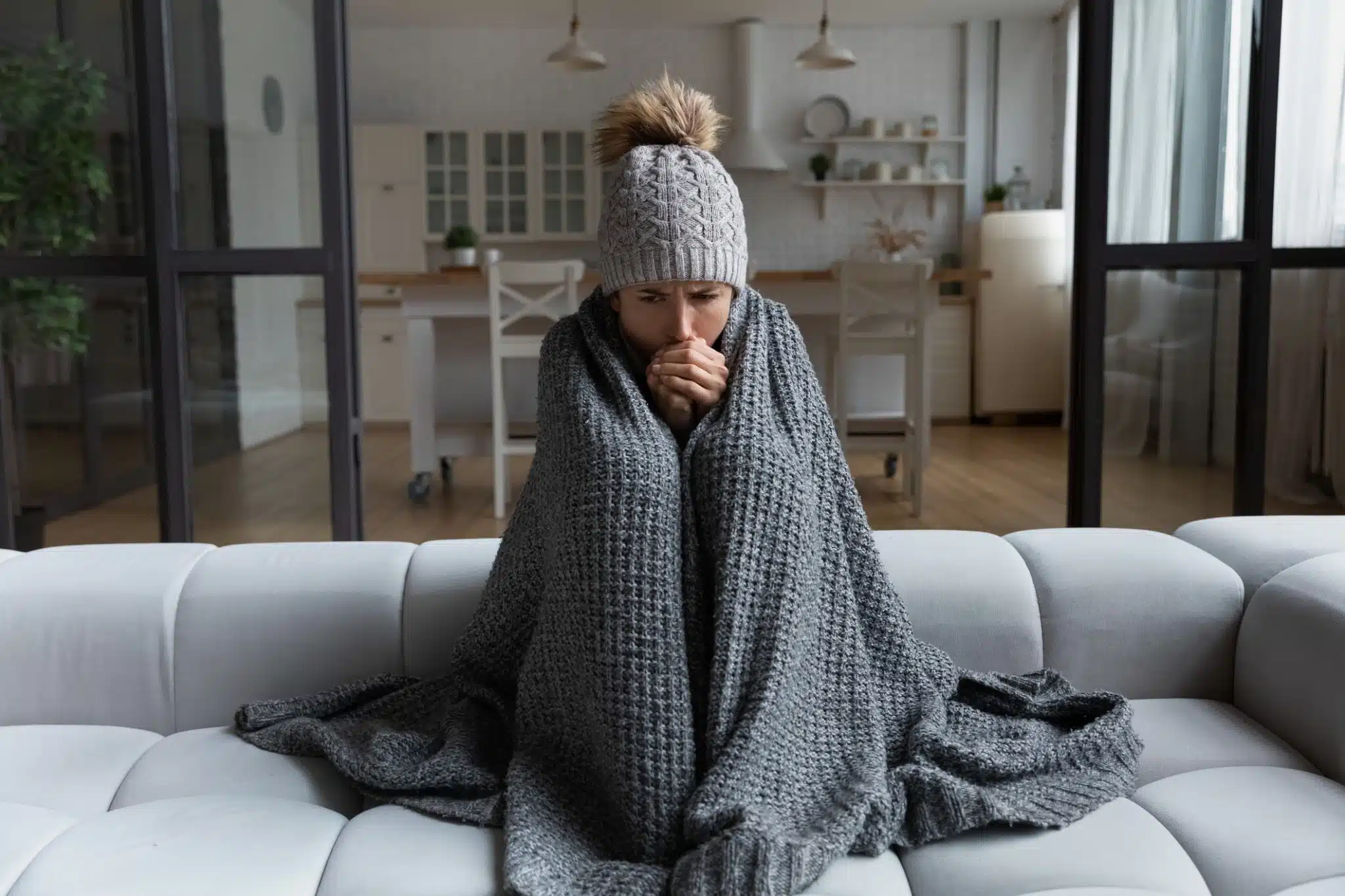A woman wrapped in a blanket on a couch attempts to stay warm after her HVAC system struggles during a winter cold snap.