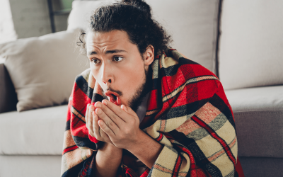 A man who is cold indoors, sits in front of the couch with a blanket on his back. He is blowing warm air on his hands because he is cold. He needs a new HVAC system from Houk Air Conditioning.