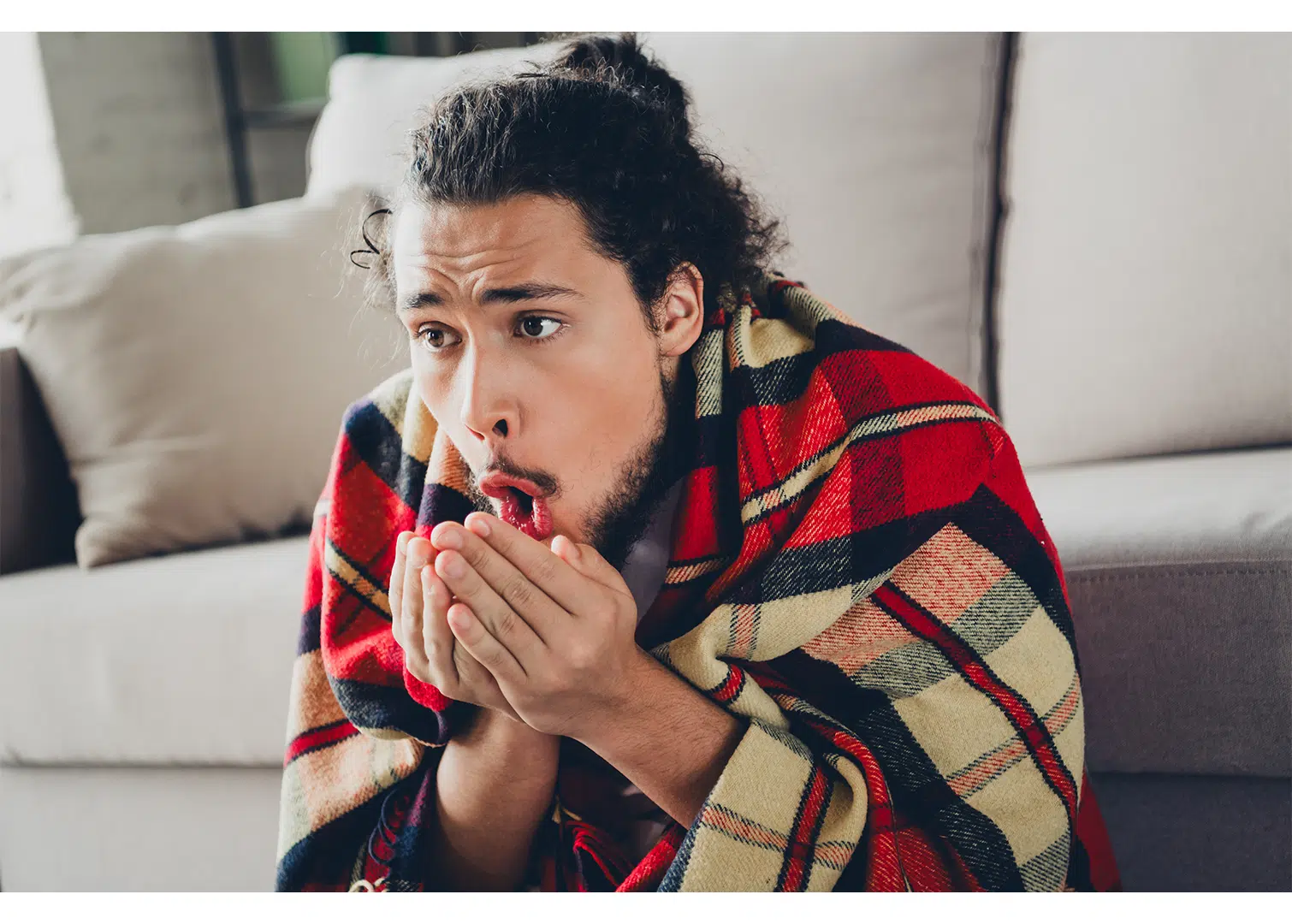 A man who is cold indoors, sits in front of the couch with a blanket on his back. He is blowing warm air on his hands because he is cold. He needs a new HVAC system from Houk Air Conditioning.