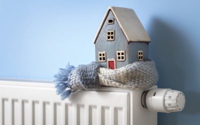 A small ceramic house wearing a knit scarf sits on top of a white radiator, symbolizing a cold and under-heated home.