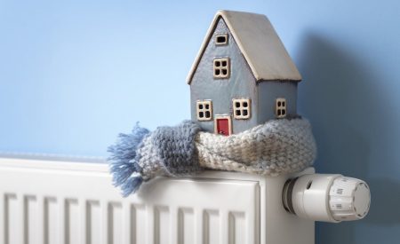 A small ceramic house wearing a knit scarf sits on top of a white radiator, symbolizing a cold and under-heated home.