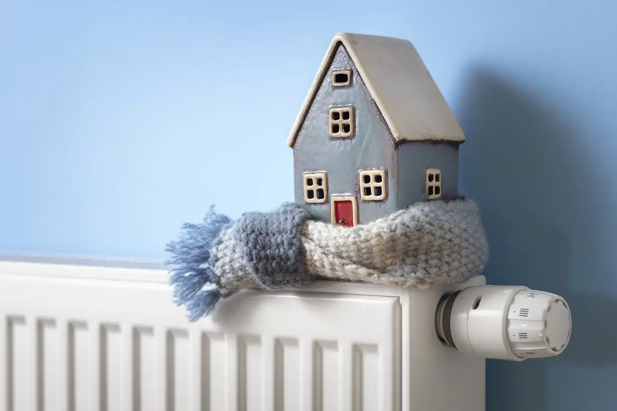 A small ceramic house wearing a knit scarf sits on top of a white radiator, symbolizing a cold and under-heated home.