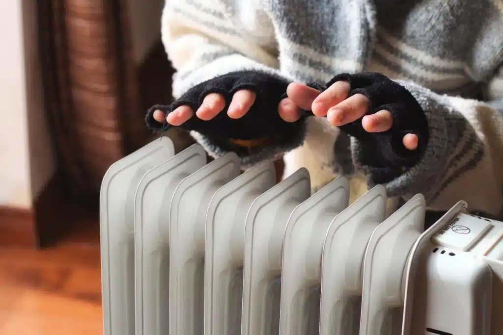 A person in a sweater and fingerless gloves warming their hands over a portable radiator heater inside a home.