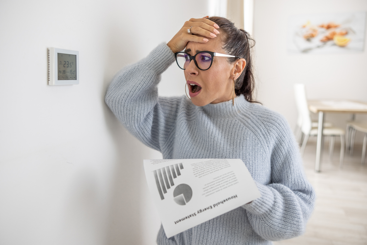 A homeowner reacting with shock to her household energy statement while standing next to her thermostat, illustrating the cost impact of AC short cycling.