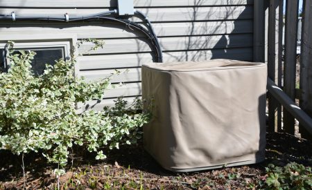 A residential AC unit still covered from winter, surrounded by budding shrubs and spring growth near a DFW home.