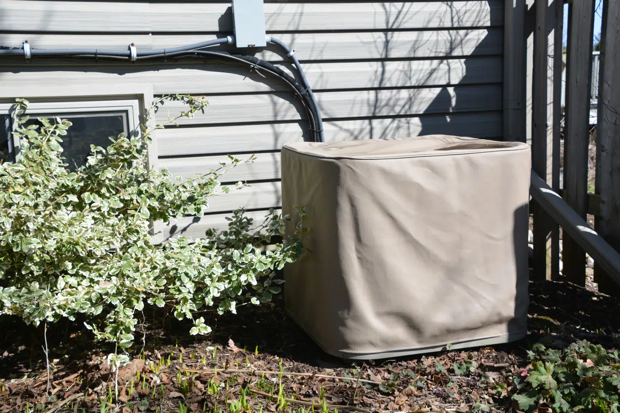 A residential AC unit still covered from winter, surrounded by budding shrubs and spring growth near a DFW home.
