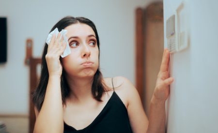 A frustrated DFW homeowner wiping sweat from her forehead while checking her thermostat as her AC cycles on and off.