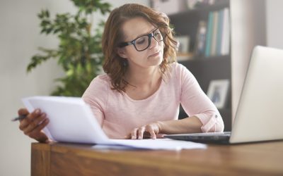 Woman in a pink top and glasses sitting at a desk, reviewing an HVAC replacement quote on paper against information on her laptop screen.