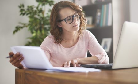 Woman in a pink top and glasses sitting at a desk, reviewing an HVAC replacement quote on paper against information on her laptop screen.