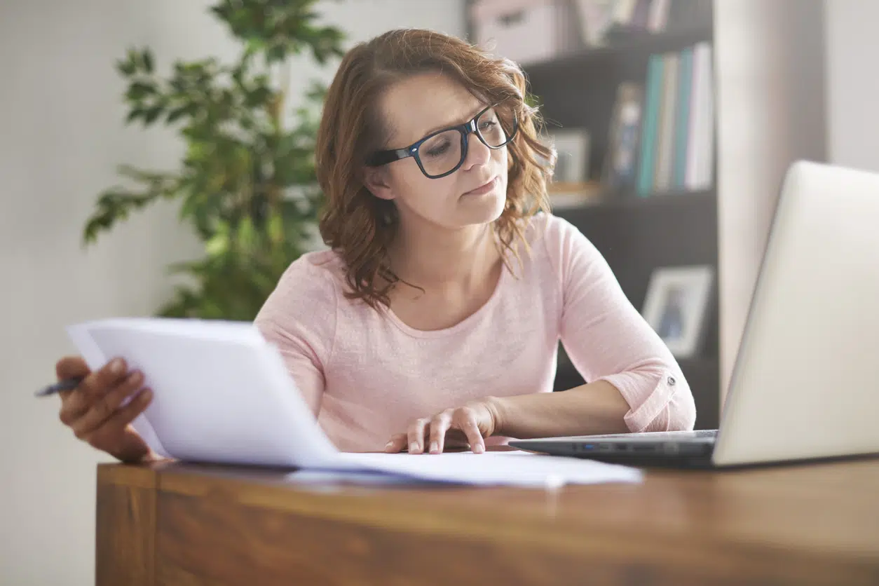 Woman in a pink top and glasses sitting at a desk, reviewing an HVAC replacement quote on paper against information on her laptop screen.