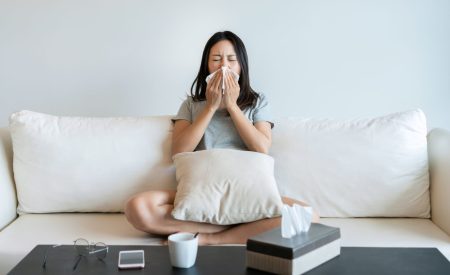 Woman sneezing into a tissue while sitting on a couch in her DFW home during spring allergy season.