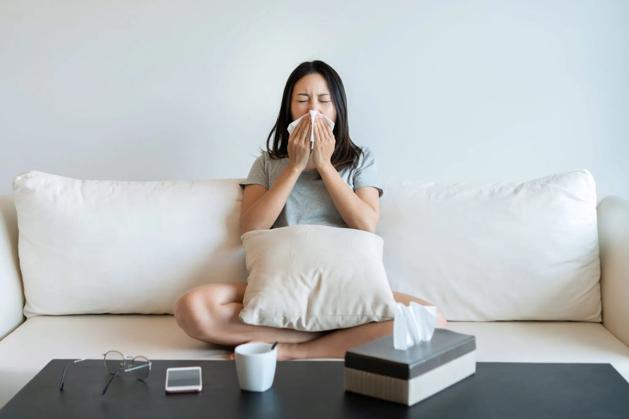 Woman sneezing into a tissue while sitting on a couch in her DFW home during spring allergy season.
