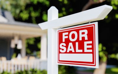 A red 'For Sale' real estate sign posted in front of a house with a white picket fence.