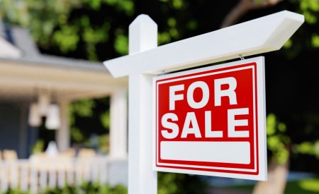A red 'For Sale' real estate sign posted in front of a house with a white picket fence.