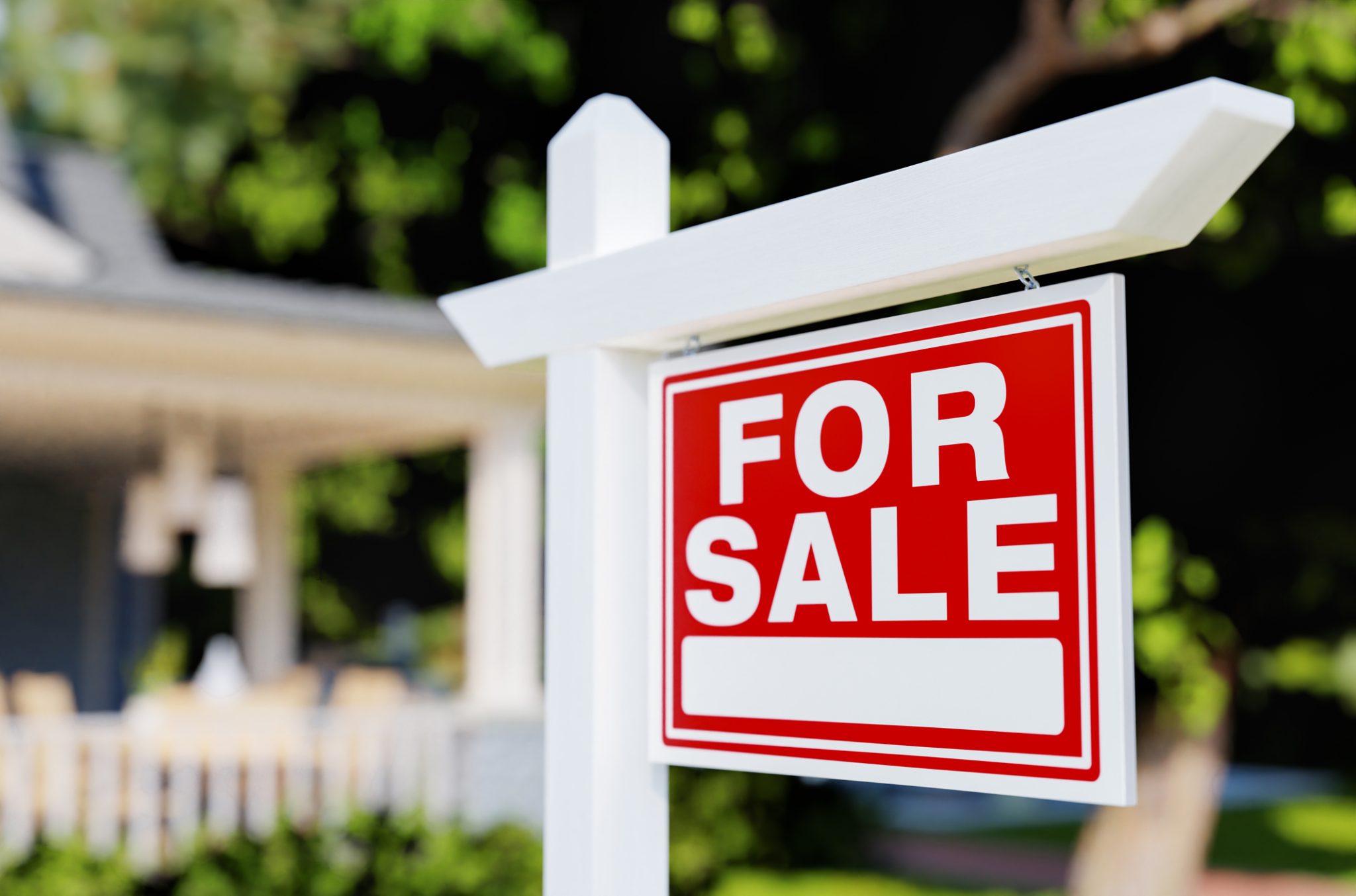 A red 'For Sale' real estate sign posted in front of a house with a white picket fence.